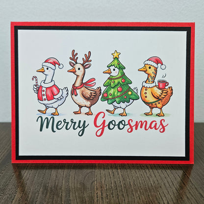 A square handmade greeting card with a red border, featuring four festive cartoon goslings dressed in Christmas attire on a white background, with the text 'Merry Goosmas' below. The card is displayed standing upright on a dark wooden table against a light gray wall.