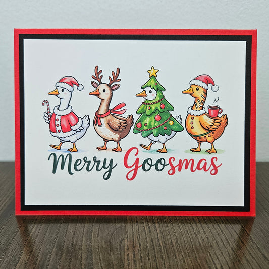 A square handmade greeting card with a red border, featuring four festive cartoon goslings dressed in Christmas attire on a white background, with the text 'Merry Goosmas' below. The card is displayed standing upright on a dark wooden table against a light gray wall.