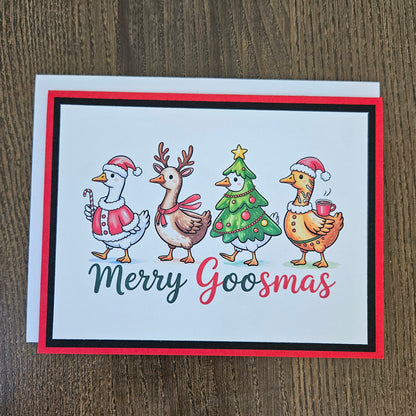 A square Christmas greeting card with a white background and colorful illustration of four festive birds around a decorated tree, with the text 'Merry Goosmas' below. The card is photographed on a brown wood surface with a white envelope behind it.