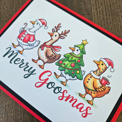 A square greeting card featuring four festive cartoon birds around Christmas trees and a reindeer, with the text 'Merry Goosmas' in black and red script. The card has a white center, a red border, and is photographed on a dark wood surface.