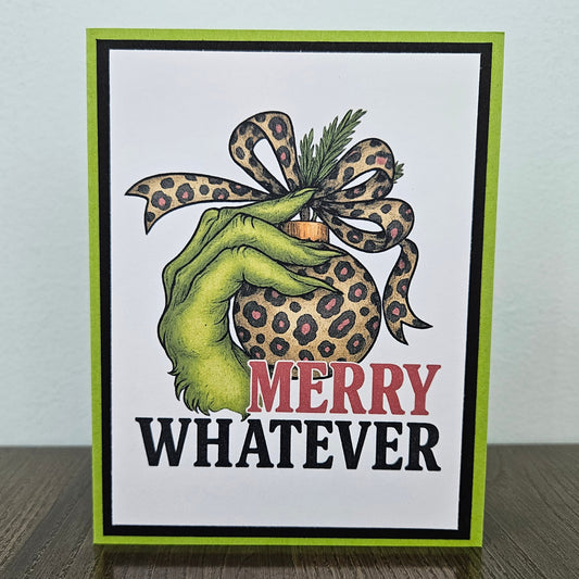 A handmade greeting card featuring a green hand holding a leopard-print ornament with a matching leopard-print bow, above the text 'MERRY WHATEVER' in red and black. The card has a white center, a black inner border, and a bright lime green outer border, standing on a dark wood surface against a light gray wall.