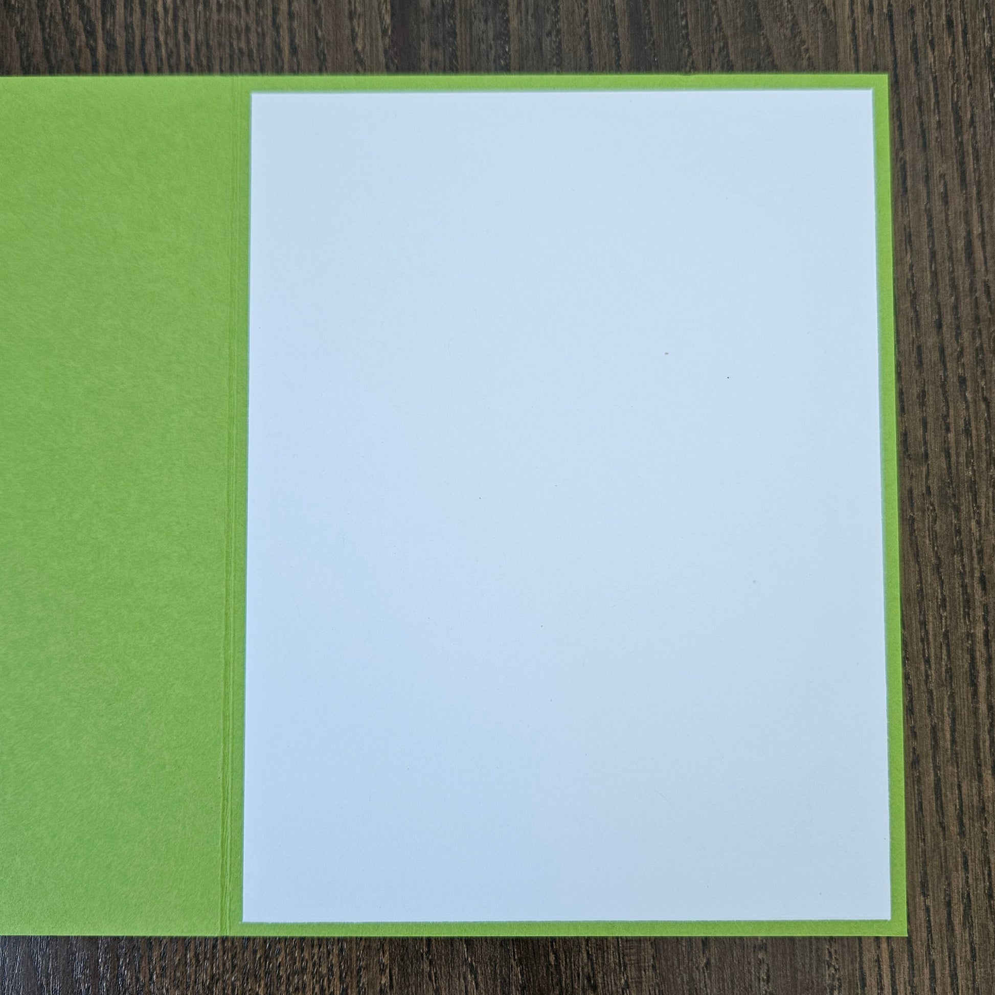 A square greeting card with a bright lime green cover and a blank white insert page, shown open on a dark wood background.