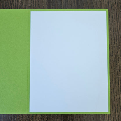 A square greeting card with a bright lime green cover and a blank white insert page, shown open on a dark wood background.