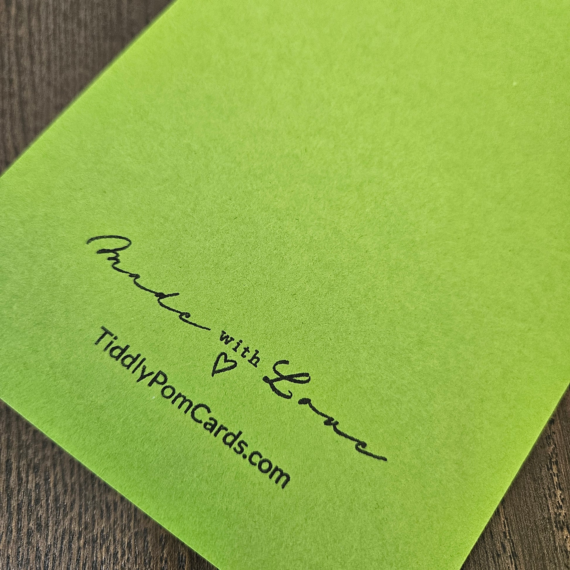 A lime green cardstock sheet with black printed text reading 'Made with Love' and 'TiddlyPomCards.com' resting on a dark wood surface.