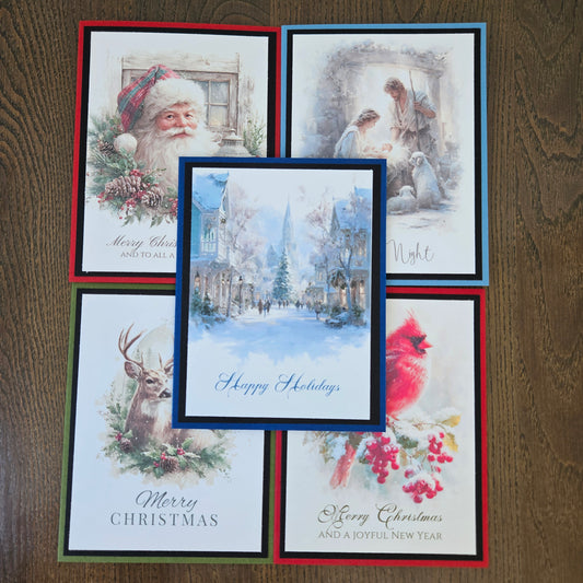 Five Christmas greeting cards arranged on a dark wood surface, featuring holiday illustrations including Santa Claus, a snowy village scene, a deer, a cardinal, and a nativity scene.