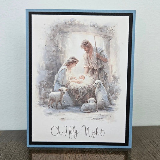 A square greeting card featuring a watercolor-style nativity scene with Mary, Joseph, baby Jesus, and sheep, bordered in light blue and black, standing on a wooden surface against a light wall.