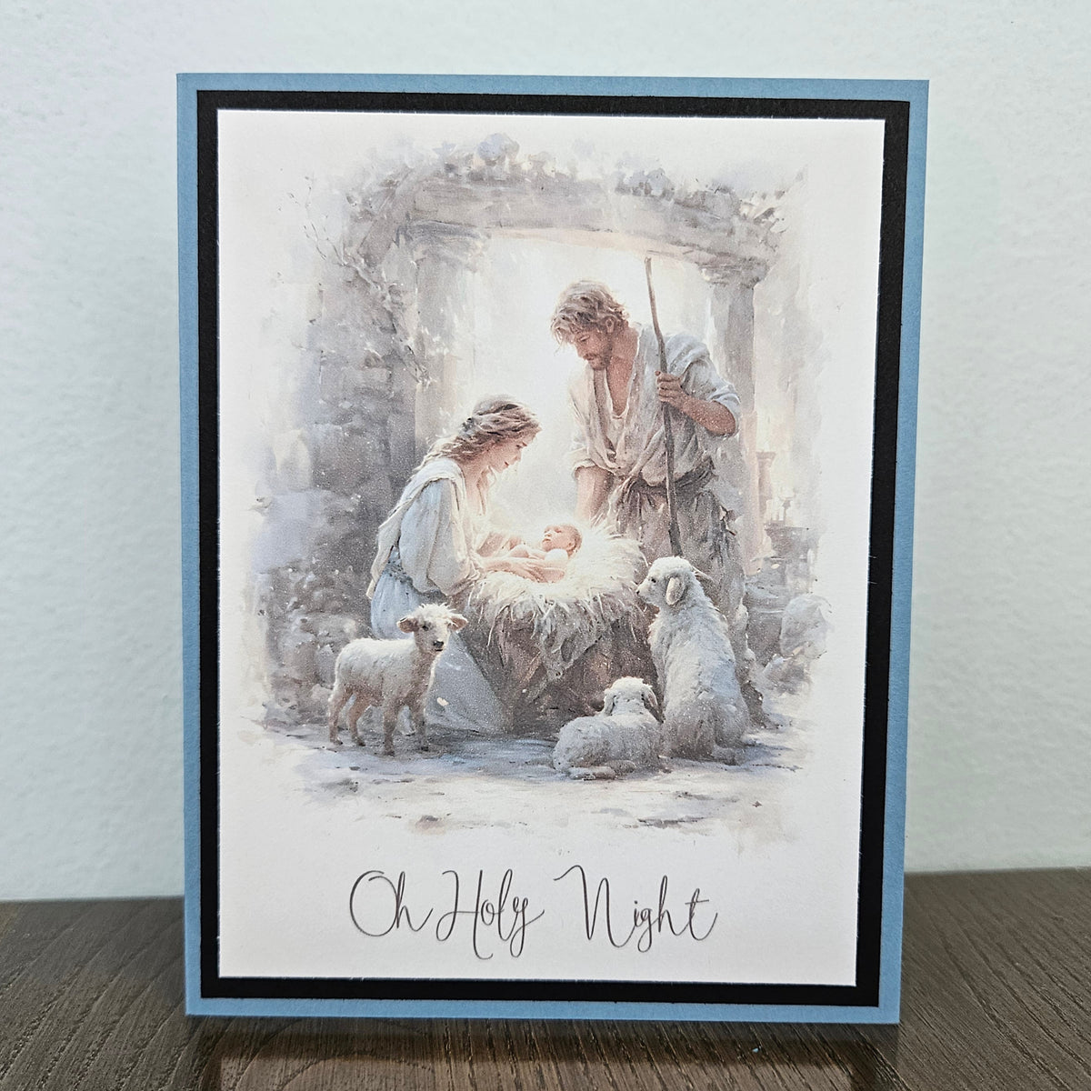 A square greeting card featuring a watercolor-style nativity scene with Mary, Joseph, baby Jesus, and sheep, bordered in light blue and black, standing on a wooden surface against a light wall.