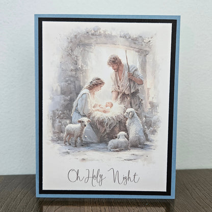 A square greeting card featuring a watercolor-style nativity scene with Mary, Joseph, baby Jesus, and sheep, bordered in light blue and black, standing on a wooden surface against a light wall.