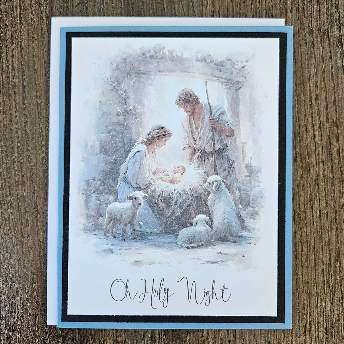 A square greeting card featuring a watercolor-style Christmas nativity scene with Mary, Joseph, baby Jesus, and sheep, placed on a brown wood surface. The card has a light blue border and the text 'Oh Holy Night' at the bottom.