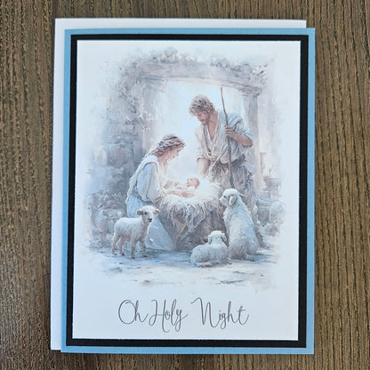 A square greeting card featuring a watercolor-style Christmas nativity scene with Mary, Joseph, baby Jesus, and sheep, placed on a brown wood surface. The card has a light blue border and the text 'Oh Holy Night' at the bottom.