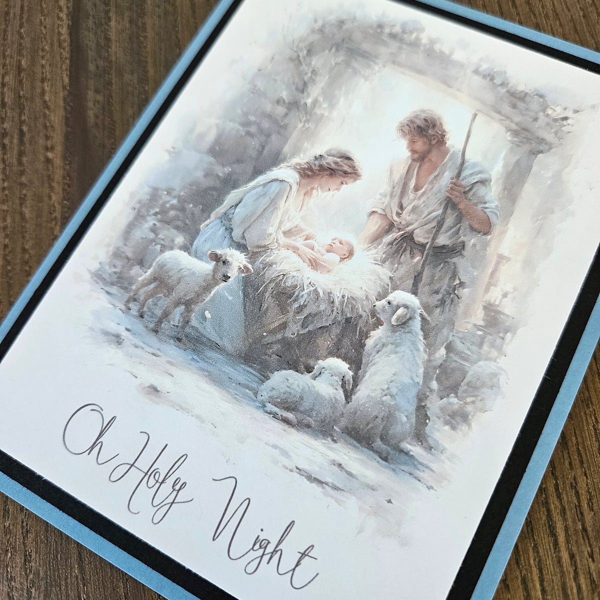 A square greeting card with a watercolor-style illustration of the Holy Family and sheep, placed on a wooden surface. The card has a light blue border and the text 'Oh Holy Night' in cursive at the bottom.