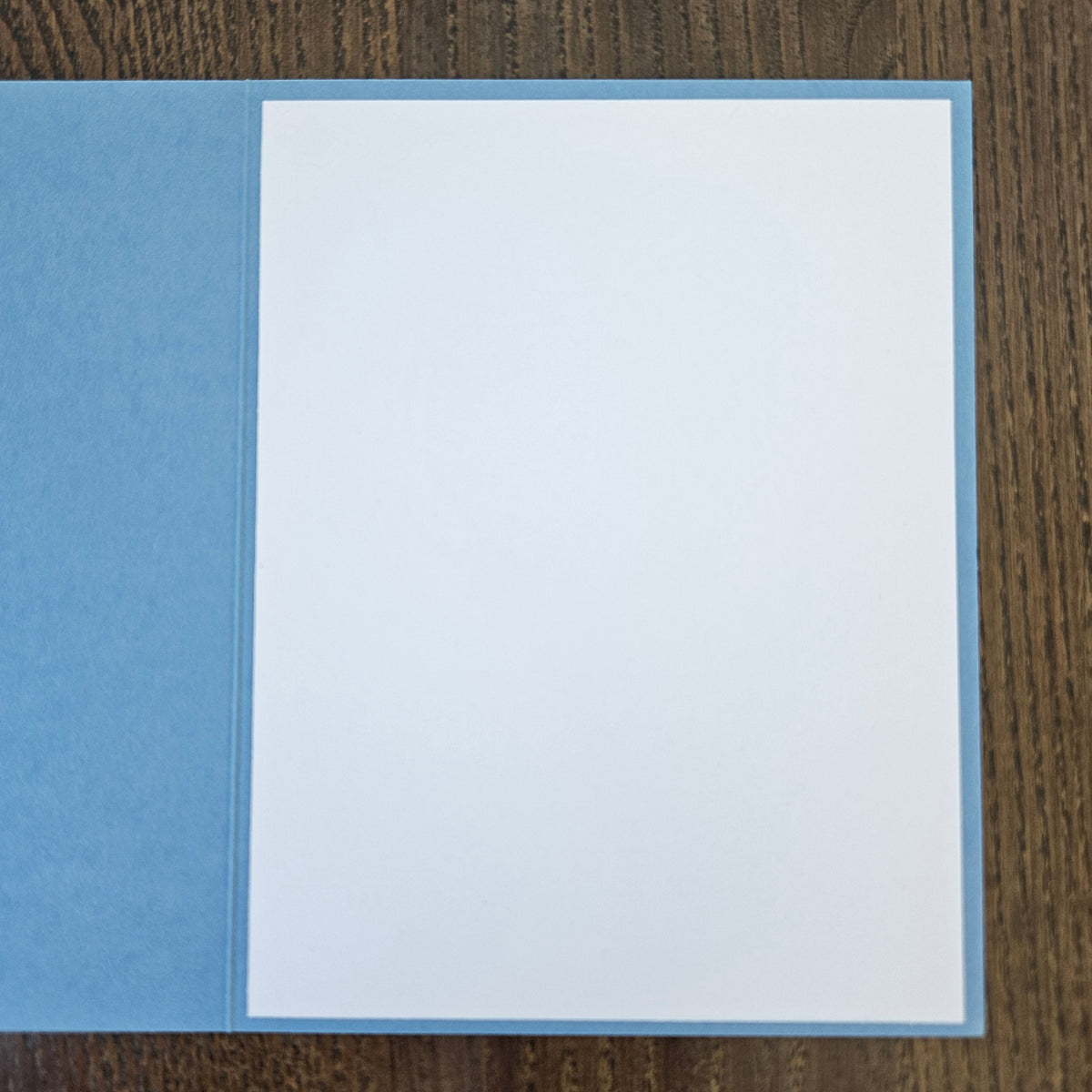 A blank white greeting card or paper sheet with a light blue border, displayed open on a dark brown wood-grain surface.