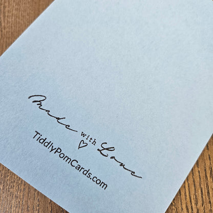 A close-up of a light blue greeting card corner printed with 'Made with Love' and 'TiddlyPomCards.com' in black script, resting on a wooden surface.