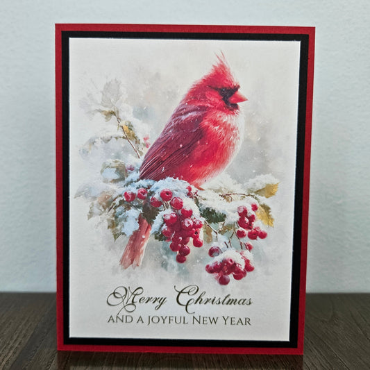 A handmade Christmas greeting card featuring a red cardinal perched on a snowy branch with red berries, bordered in red and black, with the text 'Merry Christmas AND A JOYFUL NEW YEAR' at the bottom, displayed on a dark wooden surface against a light gray wall.