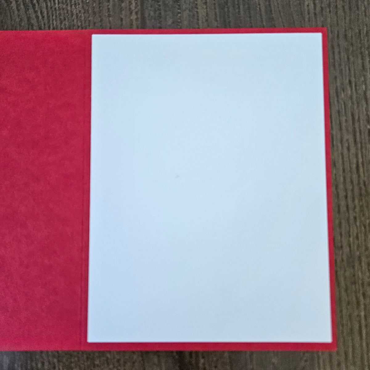 A blank white rectangular card or paper sheet placed on top of a red textured book or folder, photographed from above on a dark wood surface.
