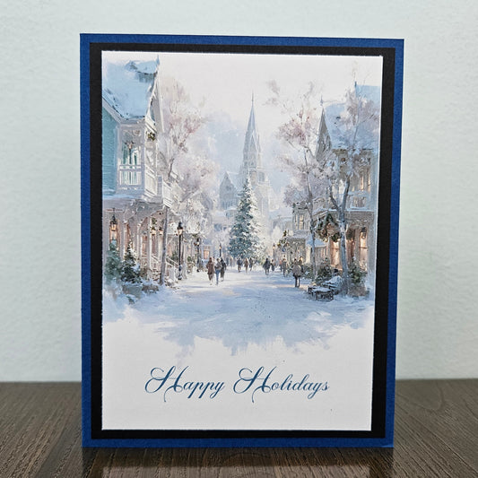 A square holiday greeting card featuring a watercolor winter street scene with snow-covered buildings, trees, and a church spire, finished with 'Happy Holidays' in blue script at the bottom. The card is displayed upright on a dark wood surface against a light gray wall.