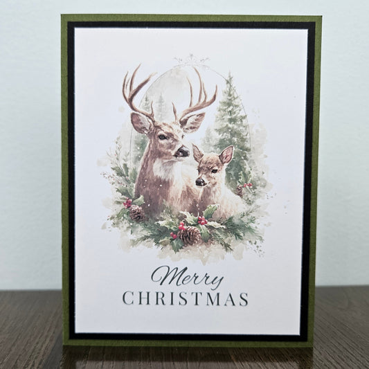 A vertical Christmas greeting card featuring an illustration of a deer and fawn with pine trees and berries, with the text 'Merry Christmas' at the bottom. The card has an olive green border and is standing on a dark wooden surface against a light gray wall.
