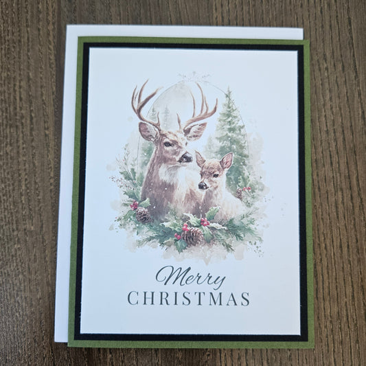 A rectangular Christmas greeting card featuring an illustration of a deer and a young fawn surrounded by evergreen branches and pinecones, with the text 'Merry CHRISTMAS' below. The card is layered over a darker green card and photographed on a brown wood surface.
