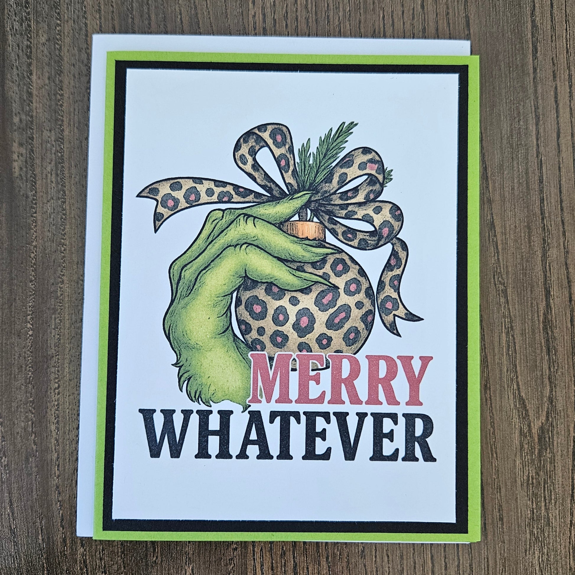 A square greeting card with a white background and a bright lime green border, featuring a hand holding a leopard-print ornament with the text 'MERRY WHATEVER' on a wooden tabletop.