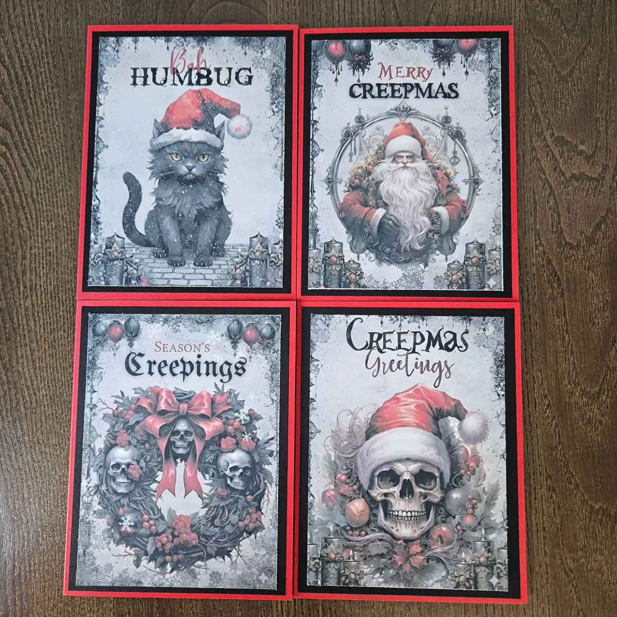 Four Christmas-themed art cards arranged in a 2x2 grid on a wooden surface. The cards feature gothic holiday illustrations: a gray cat wearing a Santa hat, Santa Claus with 'Merry Creepmas' text, two skulls in a wreath with 'Season's Creepings', and a skull wearing a Santa hat with 'Creepmas Greetings'.