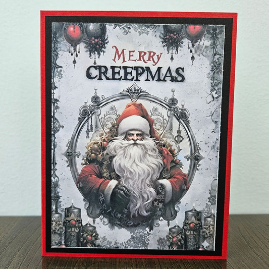 A handmade greeting card featuring a gothic Santa Claus illustration with the text 'MERRY CREEPMAS' at the top, displayed standing on a wooden surface against a light wall.