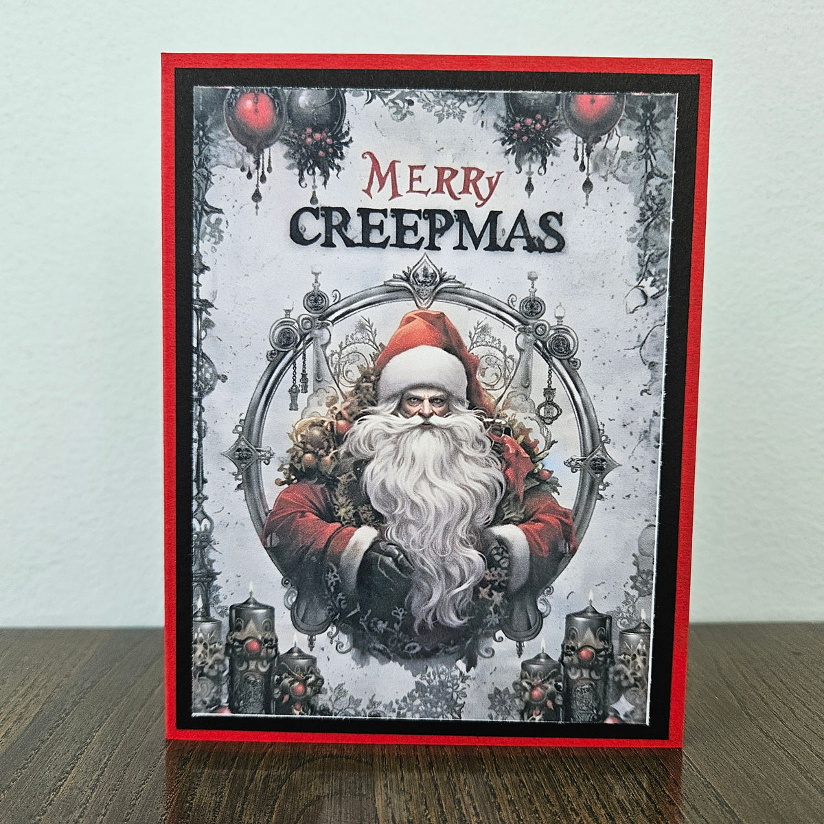 A handmade greeting card featuring a gothic Santa Claus illustration with the text 'MERRY CREEPMAS' at the top, displayed standing on a wooden surface against a light wall.