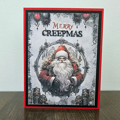 A handmade greeting card featuring a gothic Santa Claus illustration with the text 'MERRY CREEPMAS' at the top, displayed standing on a wooden surface against a light wall.