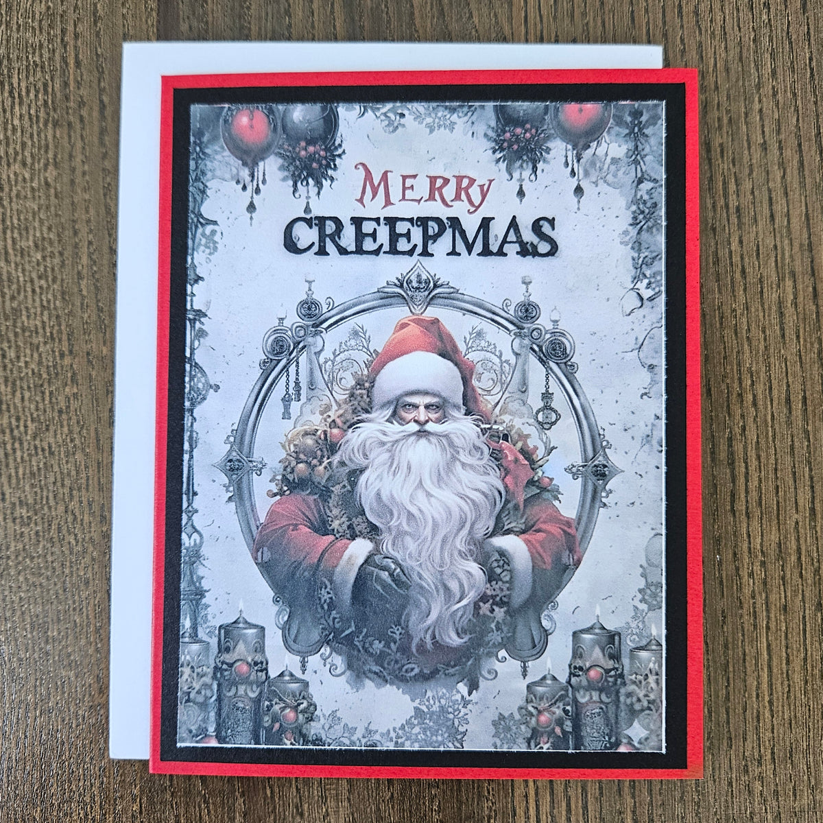 A square Christmas greeting card with a white envelope, placed on a wooden surface. The card features a gothic-style illustration of Santa Claus with a long white beard, surrounded by ornaments and candles, with the text 'MERRY CREEPMAS' at the top.