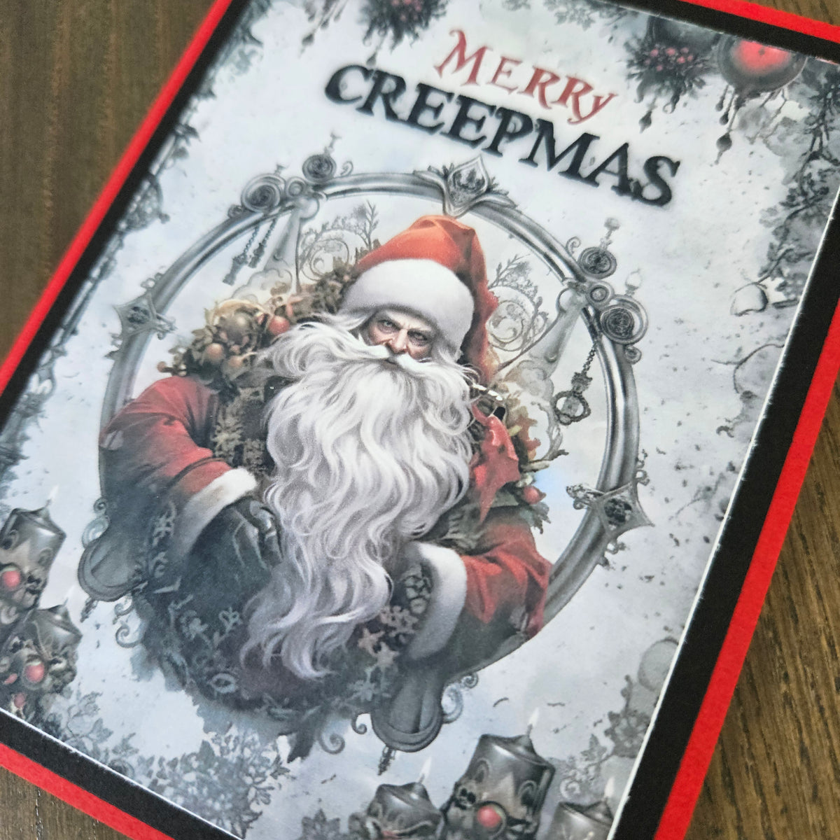 A close-up of a Christmas card featuring an illustration of Santa Claus with a long white beard, red suit, and the text 'Merry Creepmas' at the top, photographed on a wooden surface.