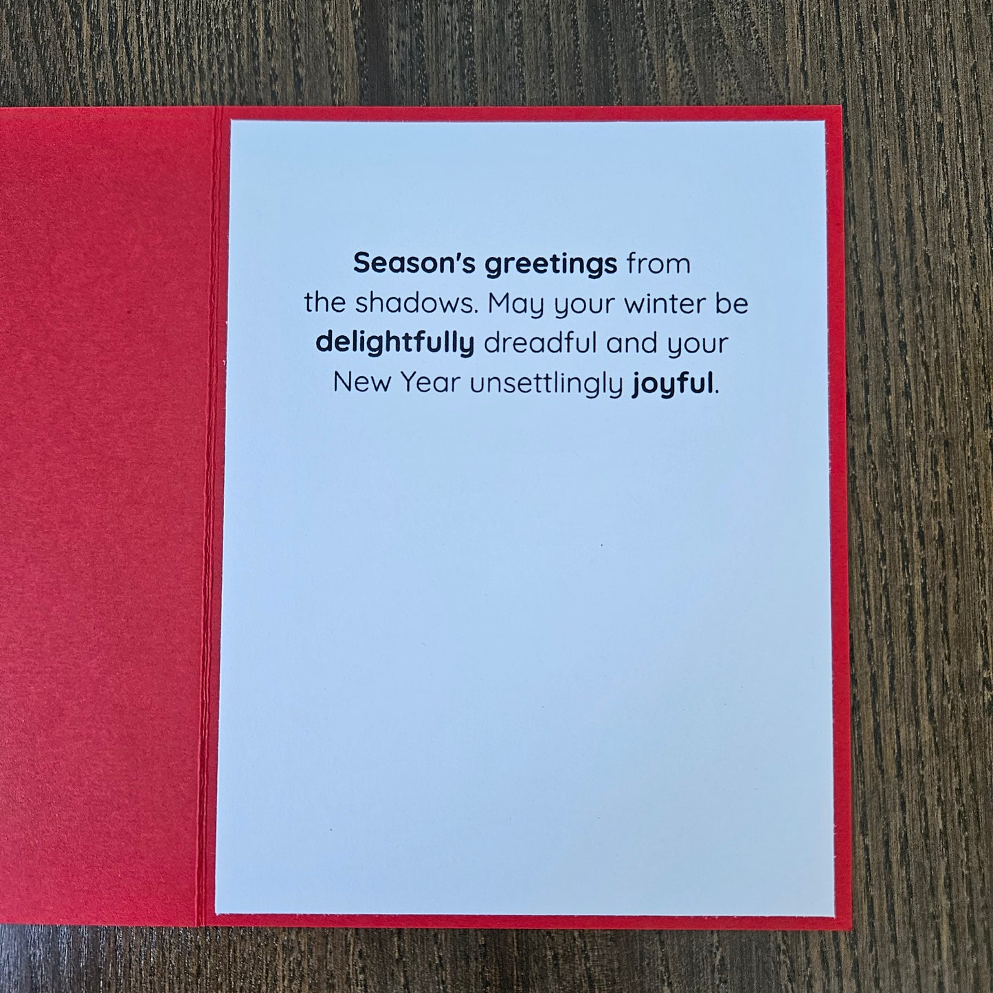 A red greeting card laid flat on a dark wood surface, showing a white interior with printed New Year's message: 'Season's greetings from the shadows. May your winter be delightfully dreadful and your New Year unsettingly joyful.