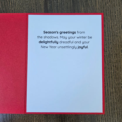 A red greeting card laid flat on a dark wood surface, showing a white interior with printed New Year's message: 'Season's greetings from the shadows. May your winter be delightfully dreadful and your New Year unsettingly joyful.