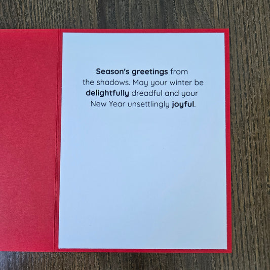 A red greeting card laid flat on a dark wood surface, showing a white interior with printed New Year's message: 'Season's greetings from the shadows. May your winter be delightfully dreadful and your New Year unsettingly joyful.