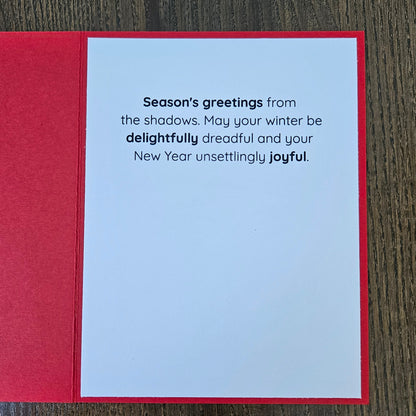 A red greeting card laid flat on a dark wood surface, showing a white interior with printed New Year's message: 'Season's greetings from the shadows. May your winter be delightfully dreadful and your New Year unsettingly joyful.