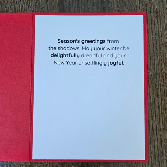 A red greeting card laid flat on a dark wood surface, showing a white interior with printed New Year's message: 'Season's greetings from the shadows. May your winter be delightfully dreadful and your New Year unsettingly joyful.