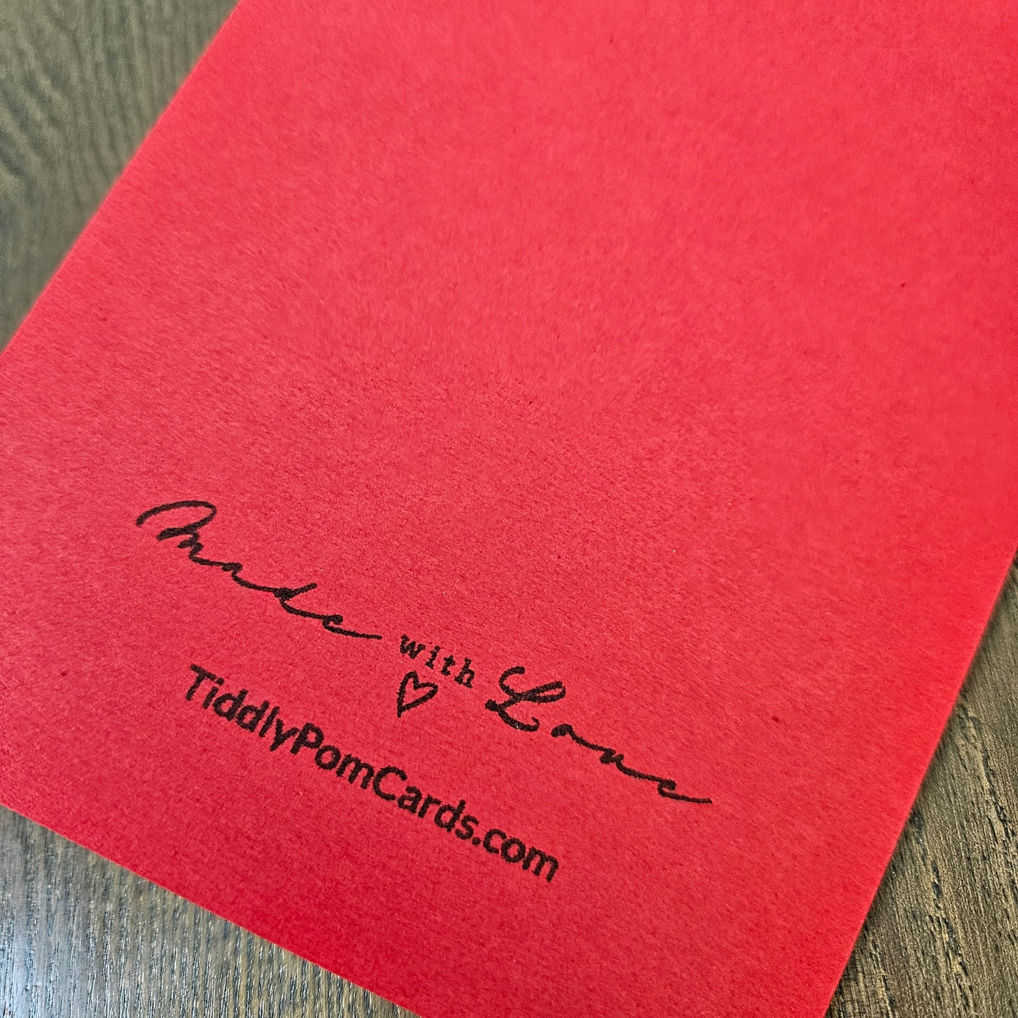 A close-up of a red textured card or paper sheet placed on a wood surface, printed with the words 'Made with Love' and 'TiddlyPomCards.com' in black script.