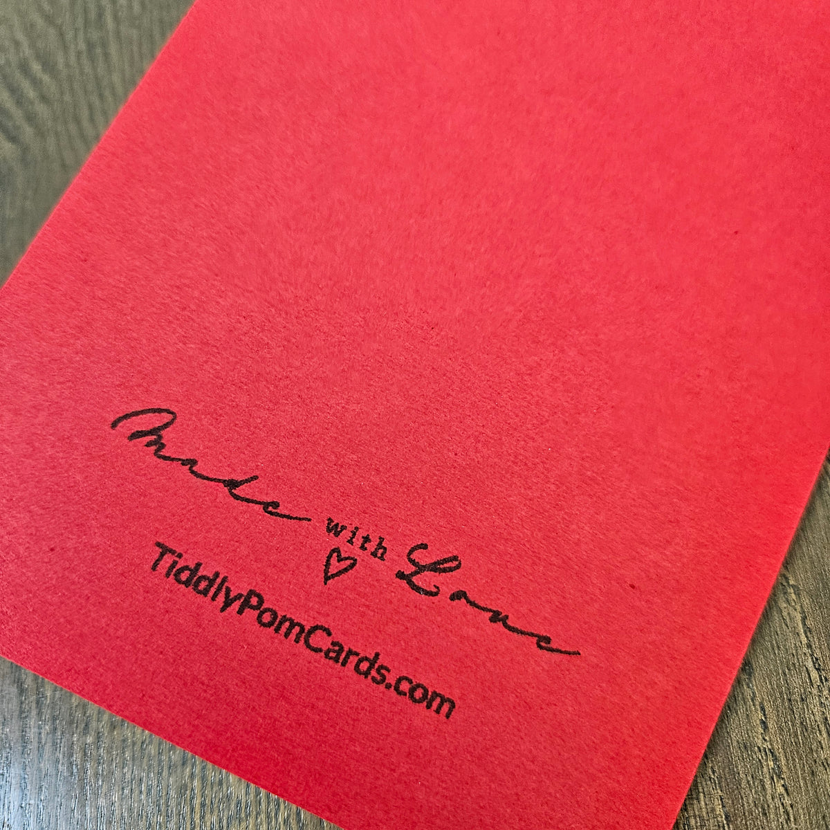 A close-up of a red textured card or paper sheet placed on a wood surface, printed with the words 'Made with Love' and 'TiddlyPomCards.com' in black script.