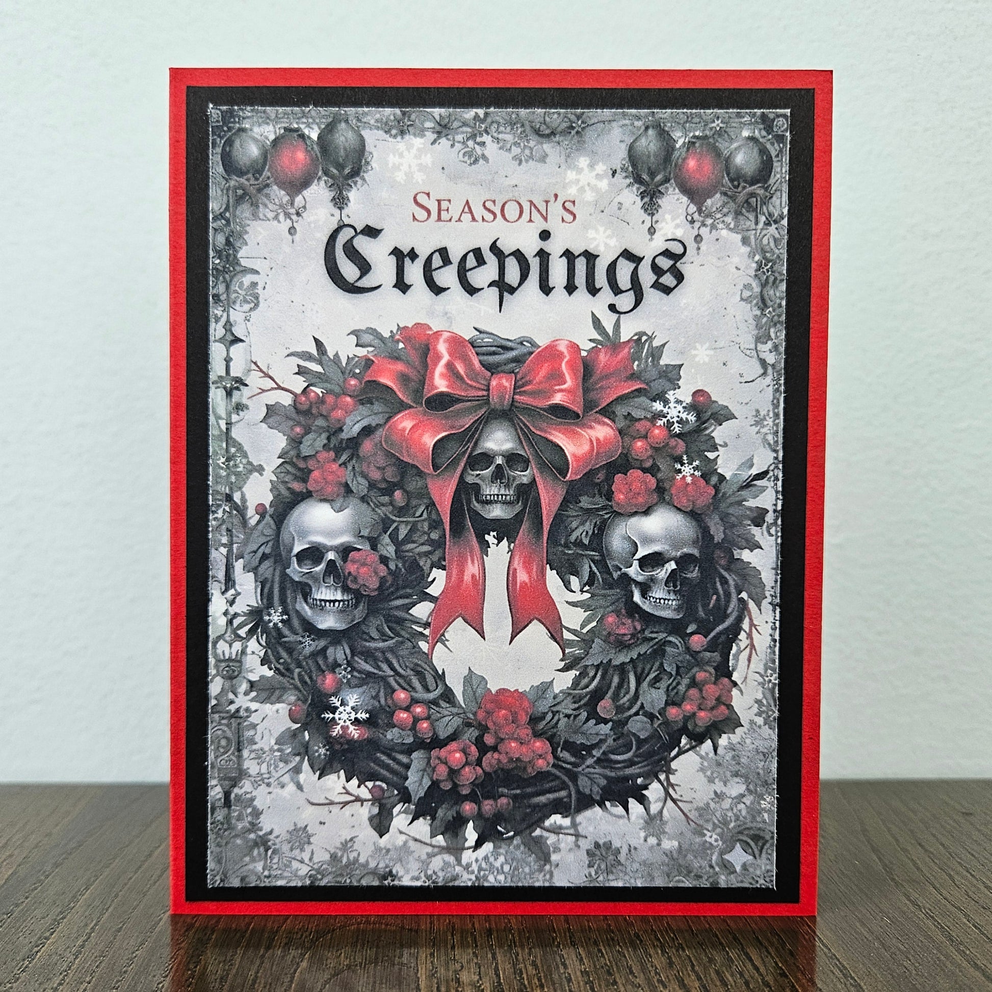 A handmade greeting card featuring a gothic skull wreath design with a large red bow, red berries, and the text 'Season's Creepings' at the top, displayed standing on a wooden surface against a light wall.
