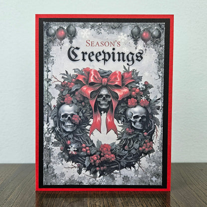 A handmade greeting card featuring a gothic skull wreath design with a large red bow, red berries, and the text 'Season's Creepings' at the top, displayed standing on a wooden surface against a light wall.