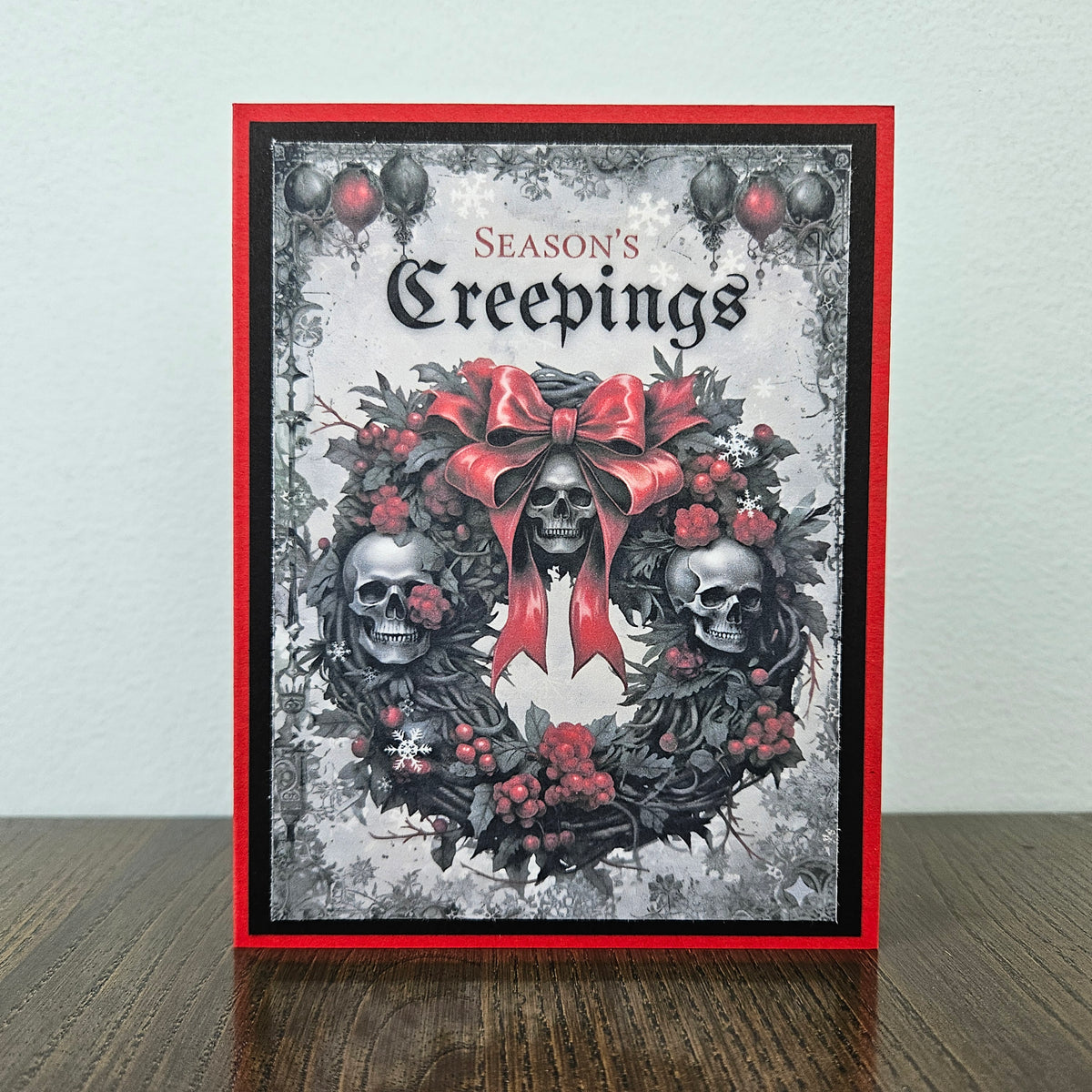 A handmade greeting card featuring a gothic skull wreath design with a large red bow, red berries, and the text 'Season's Creepings' at the top, displayed standing on a wooden surface against a light wall.