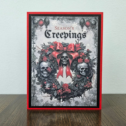 A handmade greeting card featuring a gothic skull wreath design with a large red bow, red berries, and the text 'Season's Creepings' at the top, displayed standing on a wooden surface against a light wall.