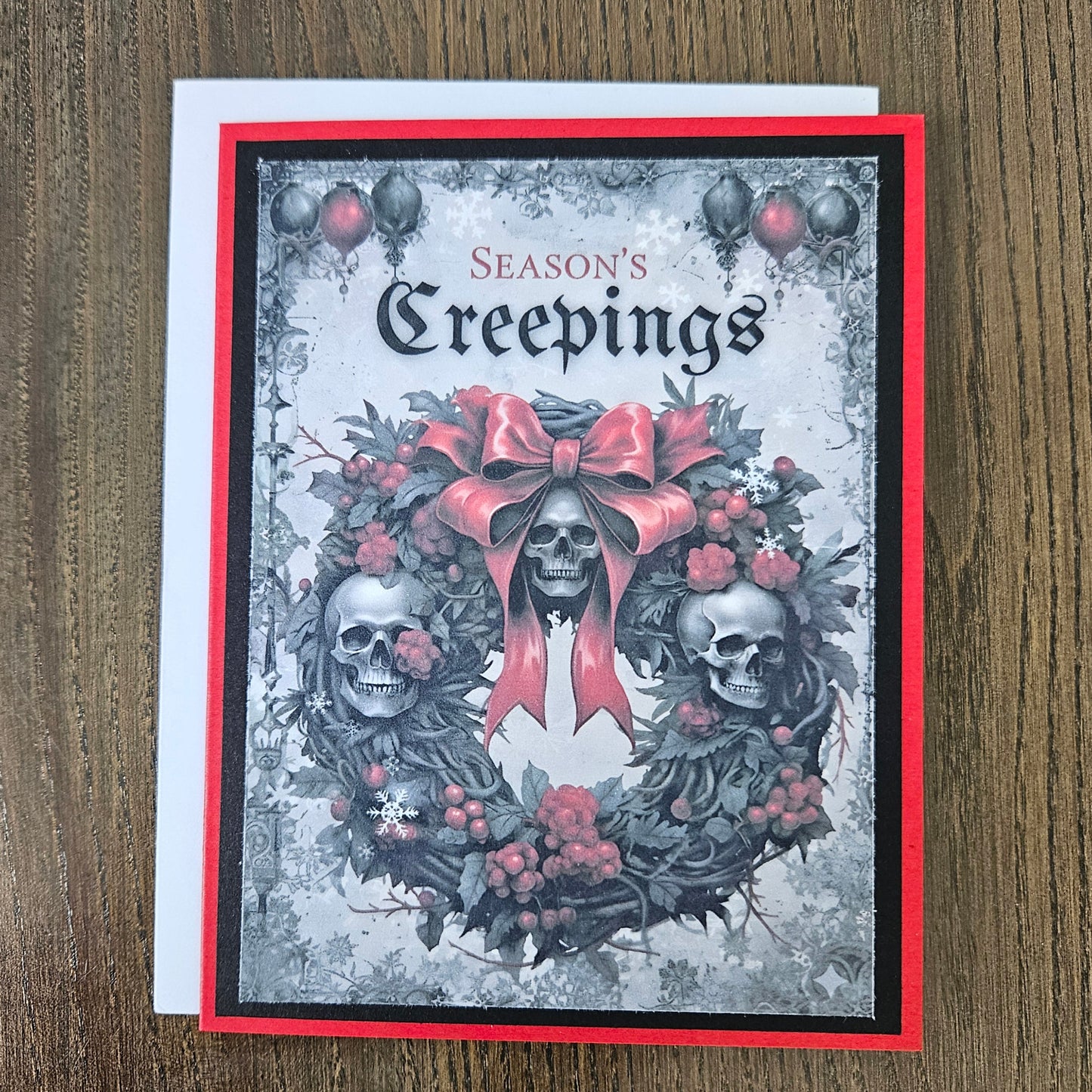 A square greeting card titled 'Season's Creepings' featuring a gothic wreath design with two skulls, red berries, and a large red bow, photographed on a wooden surface with a white envelope behind it.