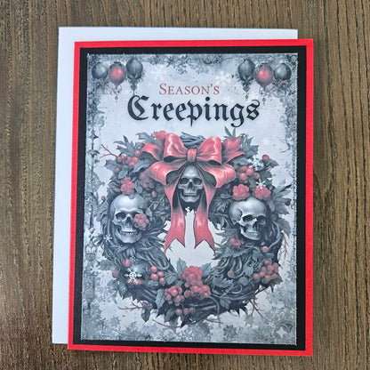 A square greeting card titled 'Season's Creepings' featuring a gothic wreath design with two skulls, red berries, and a large red bow, photographed on a wooden surface with a white envelope behind it.