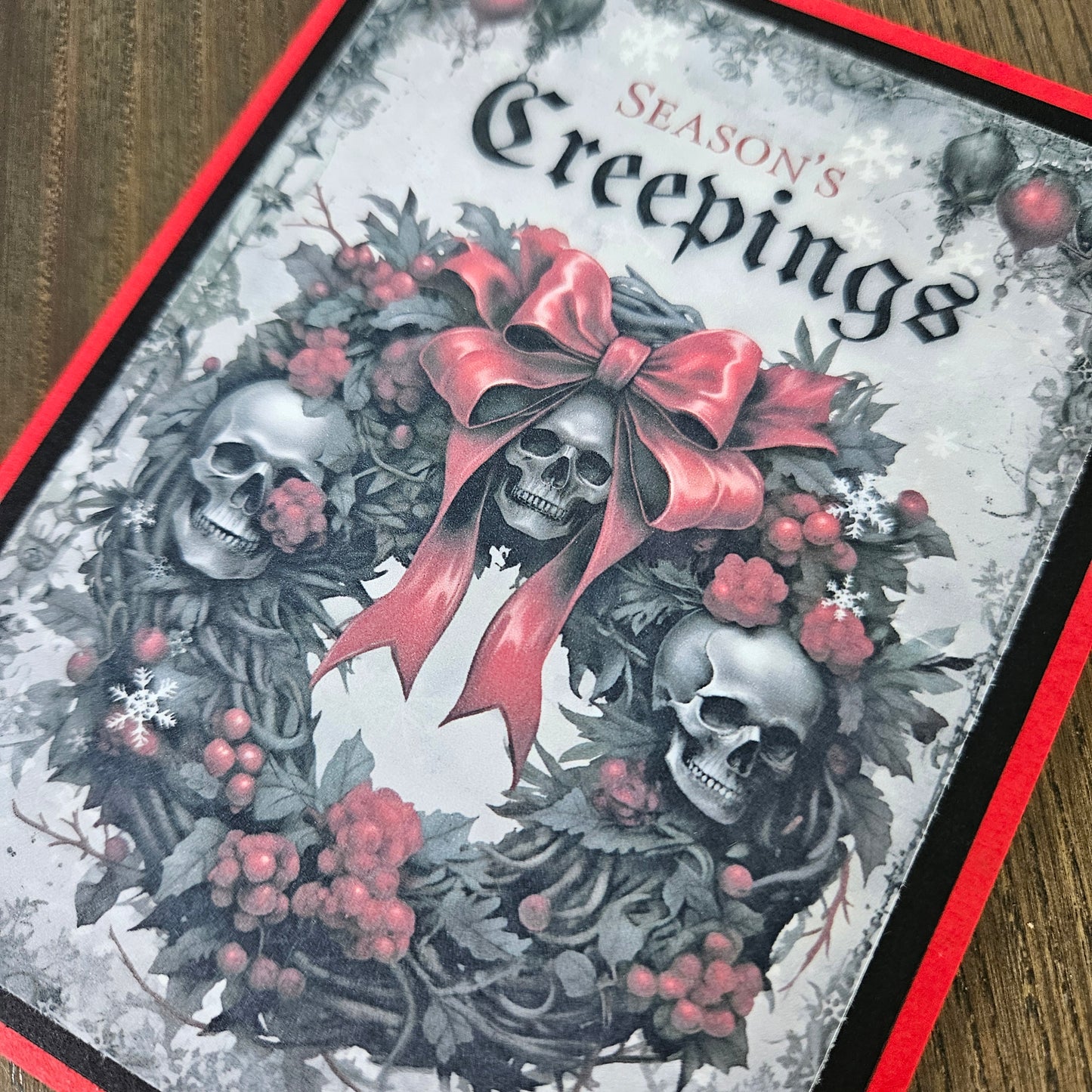 A close-up of a Christmas greeting card featuring three skulls in a wreath with a large red bow, red berries, and the text 'Season's Creepings' on a snowy gray background, bordered by red and black and photographed on a wooden surface.