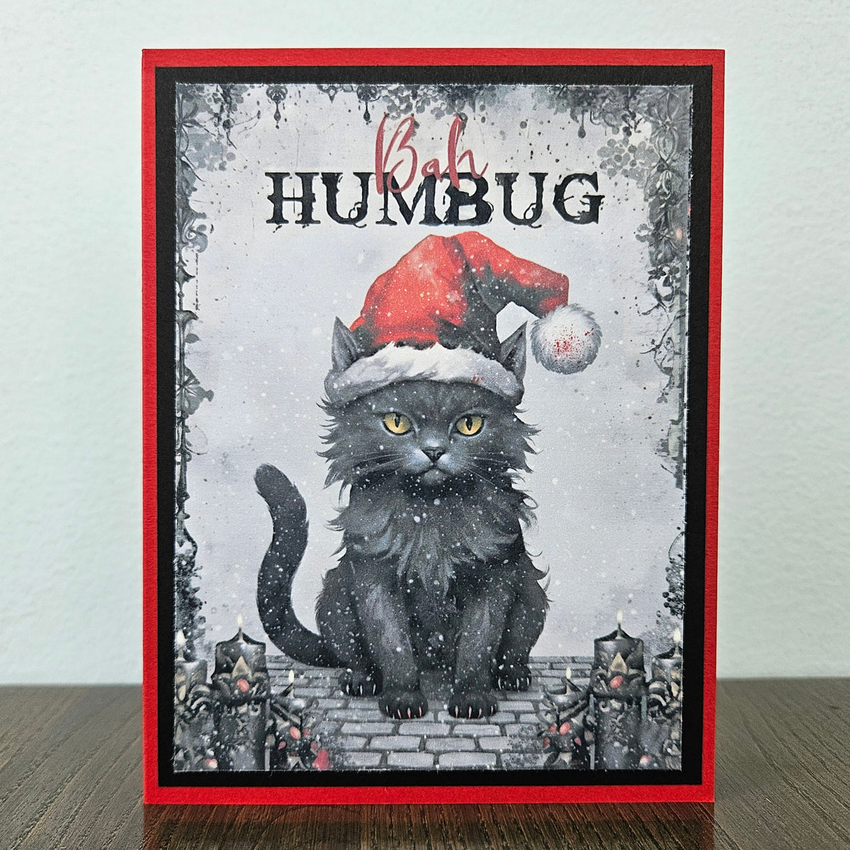 A handmade greeting card featuring a black cat wearing a red Santa hat, sitting on a brick path with the text 'Bob HUMBUG' above it. The card has a red border and is displayed standing on a dark wooden surface against a light gray wall.
