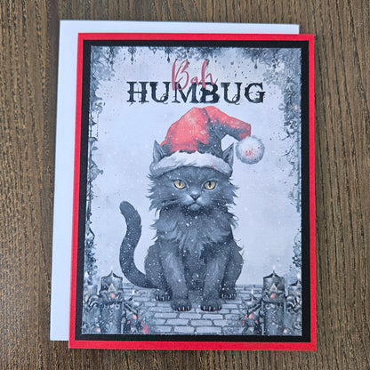 A handmade greeting card featuring a fluffy black cat wearing a red Santa hat, with the text 'Baby HUMBUG' on a rustic wood background.