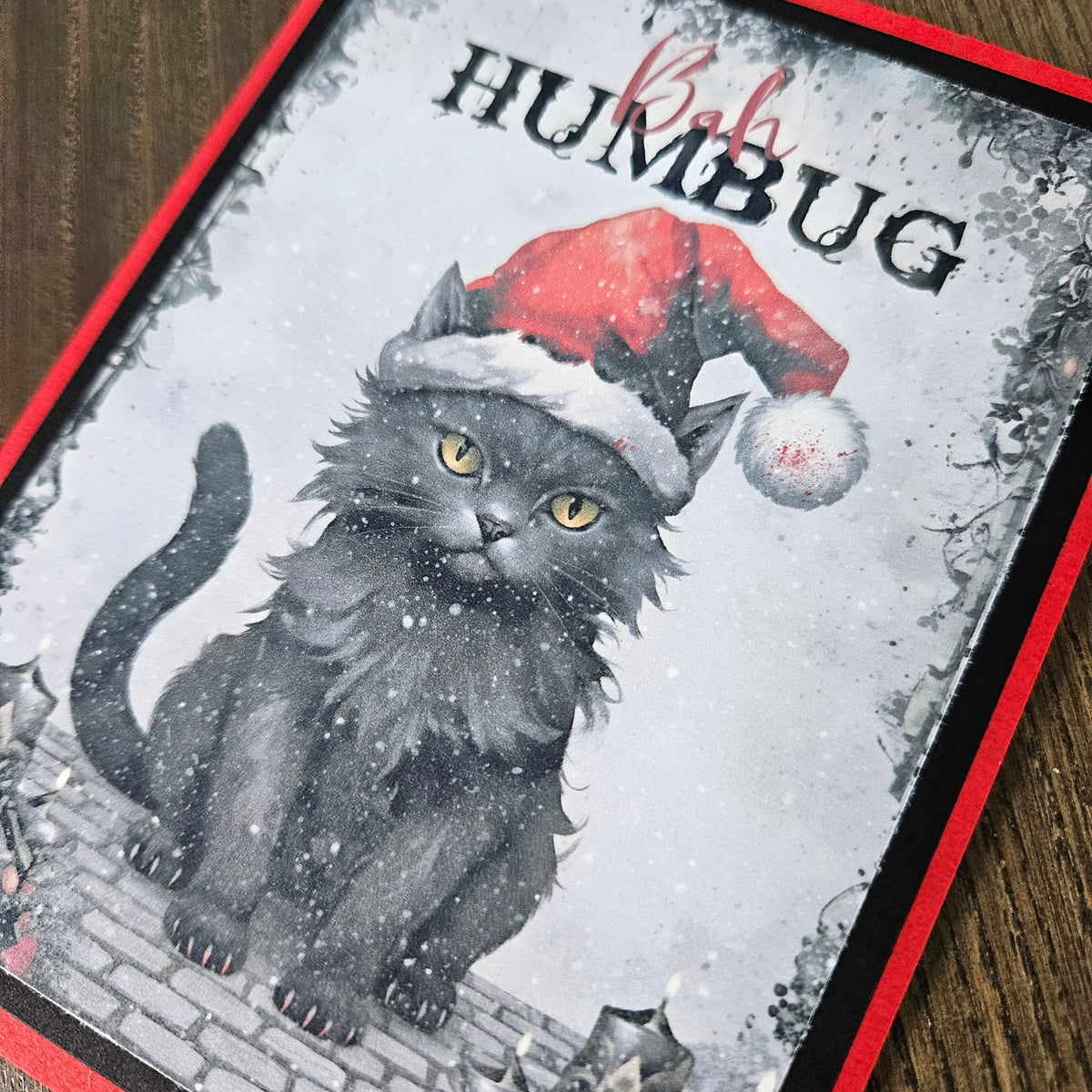A square printed card or art piece featuring a black cat wearing a red Santa hat, with the word 'HUMBUG' at the top, photographed on a wooden surface.