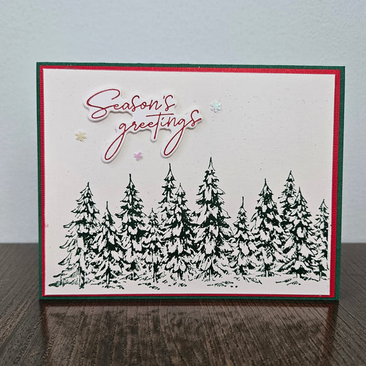 A handmade greeting card with a white background, featuring a black stamped pine tree line at the bottom, red and green border stripes, and 'Season's greetings' in red script with small pastel star accents. The card is displayed standing upright on a dark wooden surface against a light gray wall.