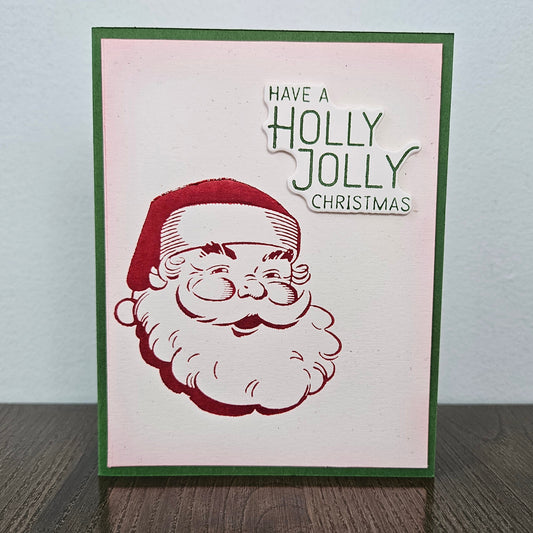 A handmade Christmas greeting card with a green border, featuring a red and white stamped illustration of Santa's face and the text 'HAVE A HOLLY JOLLY CHRISTMAS' on a light pink background, displayed on a dark wooden surface against a light gray wall.