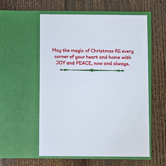 A Christmas greeting card shown open on a wooden surface, with a green card base and white insert. The insert features red printed holiday text and a small green decorative line in the center.