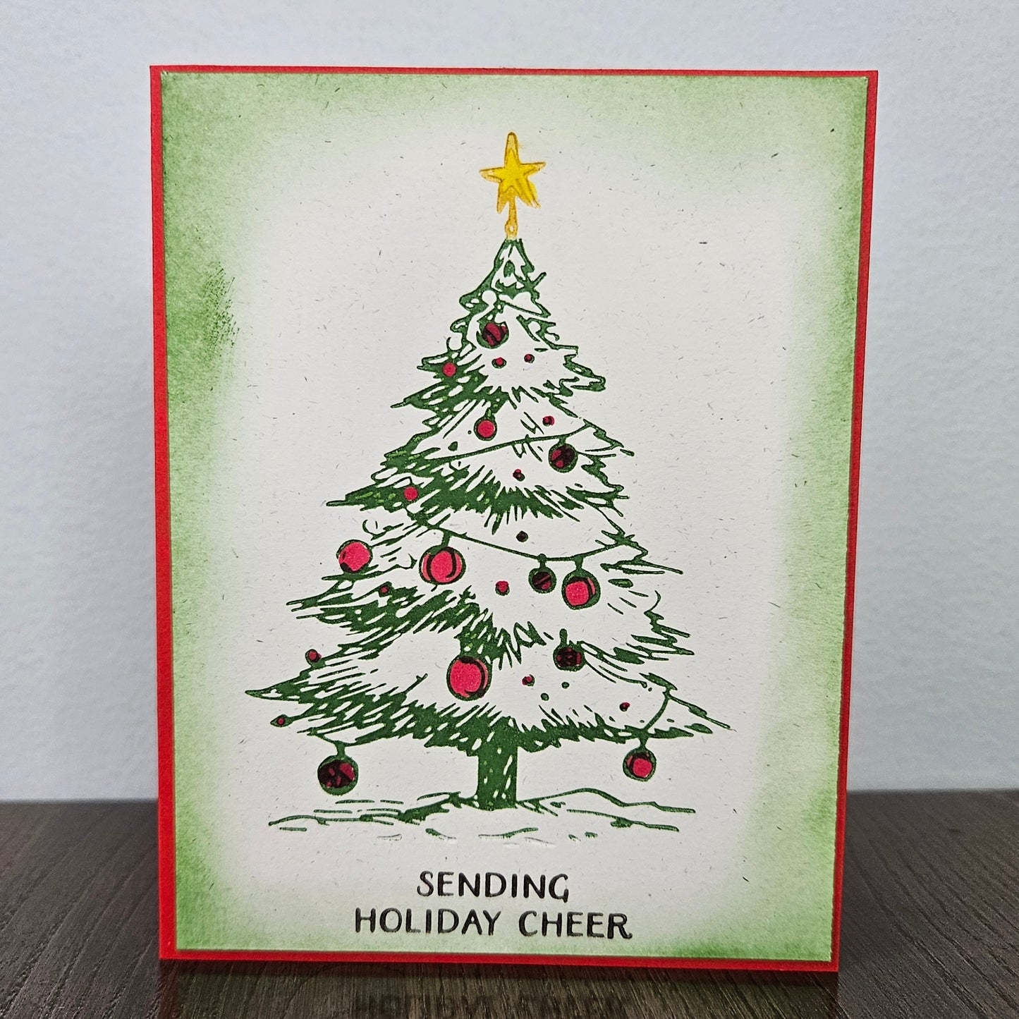 A handmade holiday greeting card featuring a green Christmas tree with red ornaments and a gold star, bordered in red with the text 'SENDING HOLIDAY CHEER' at the bottom, displayed on a dark wooden surface against a light gray wall.